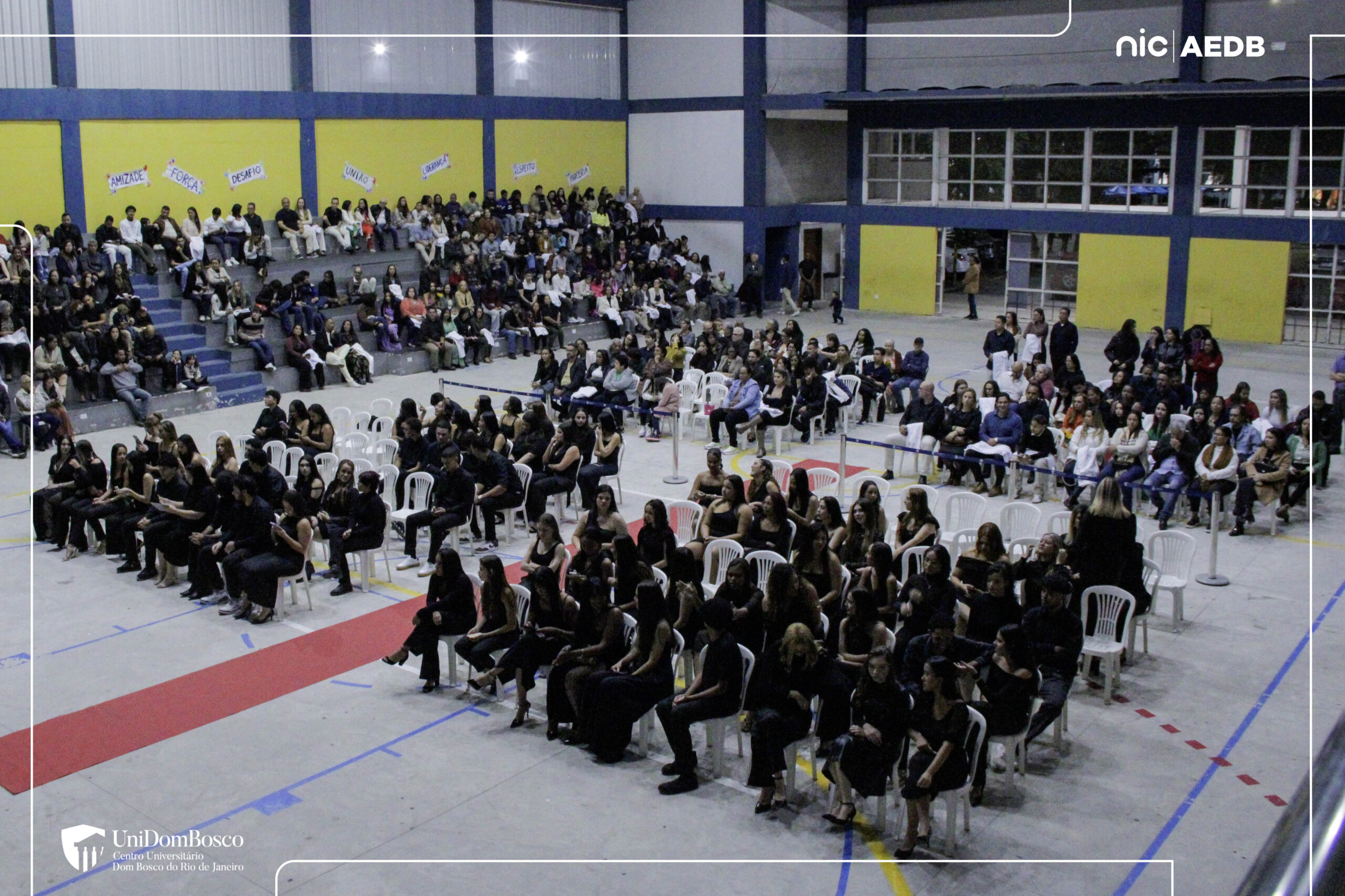 Vista aérea da Arena AEDB lotada durante a Cerimônia do Jaleco do UniDomBosco-RJ. Estudantes em roupas pretas ocupam cadeiras brancas no centro, enquanto familiares e convidados estão nas arquibancadas ao fundo. Há um tapete vermelho e decorações com palavras inspiradoras nas paredes.