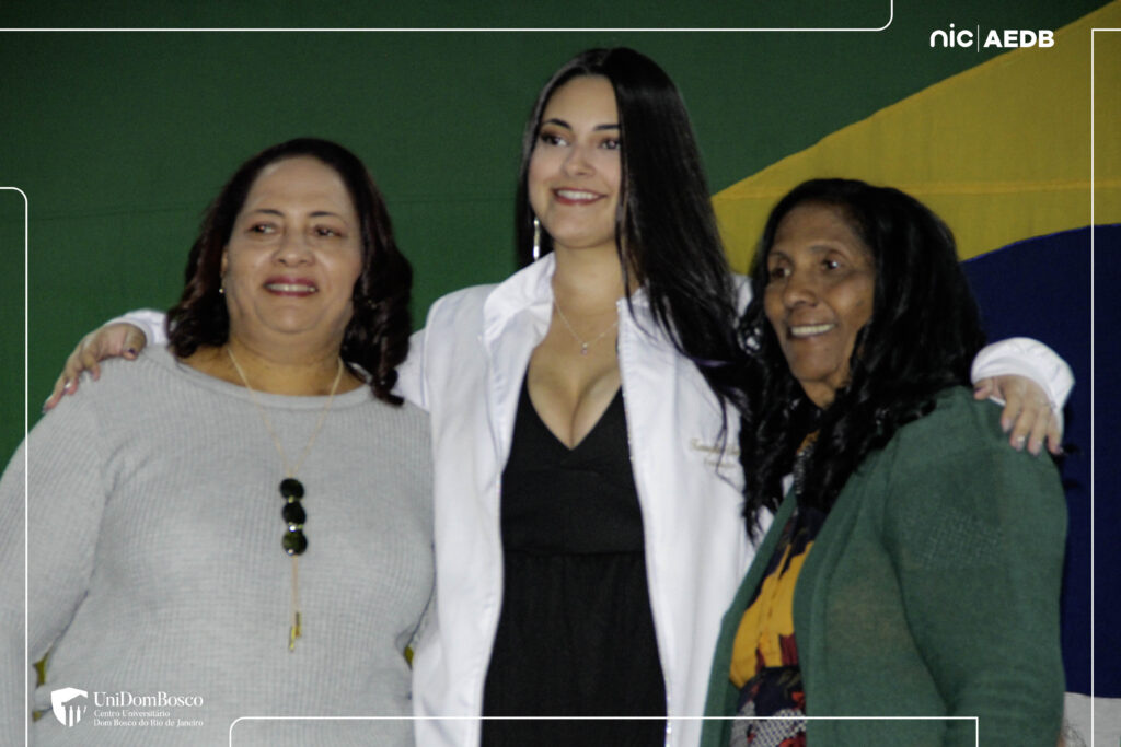 Uma jovem estudante de Farmácia, usando seu novo jaleco branco e com uma medalha, sorri abraçada a duas mulheres mais velhas (provavelmente familiares) em frente a bandeiras e um fundo verde durante a Cerimônia do Jaleco do UniDomBosco-RJ.