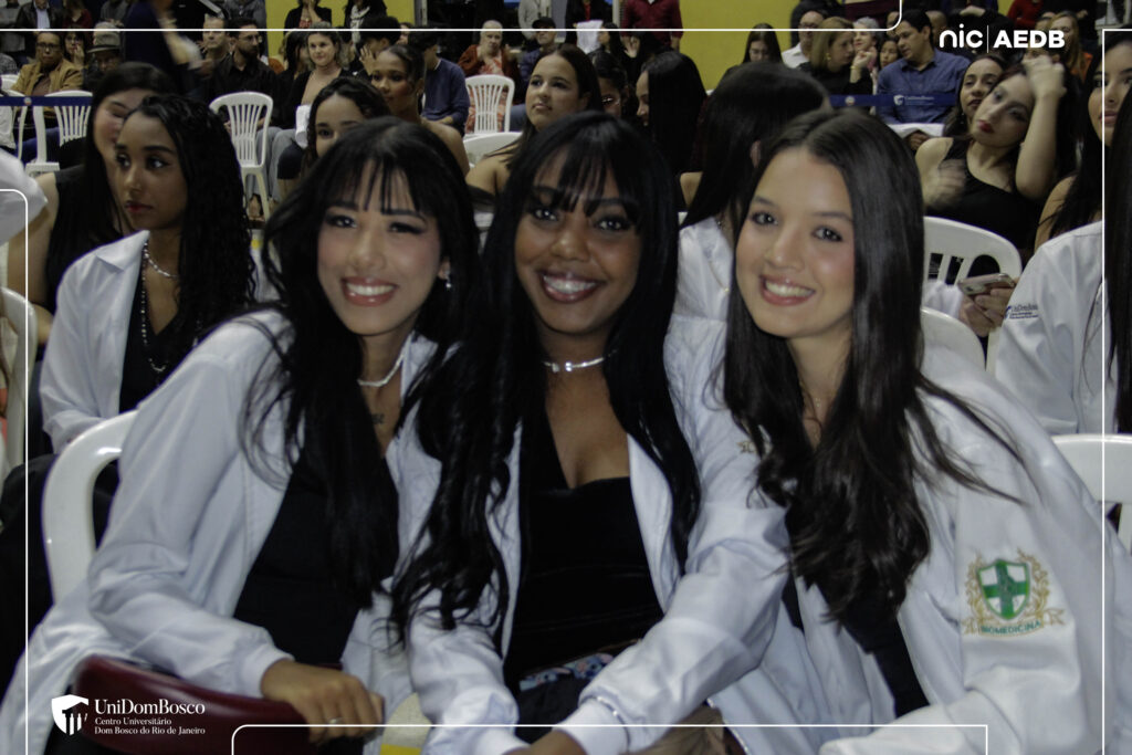 Três estudantes de cursos da saúde do UniDomBosco-RJ, usando jalecos brancos sobre roupas pretas, sorrindo e posando juntos durante a Cerimônia do Jaleco na Arena AEDB. Uma delas tem o logo do curso de Biomedicina no braço.